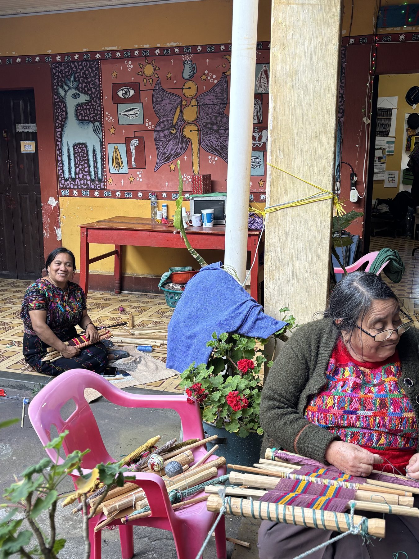 Inside Trama Textiles — two weavers at work in a courtyard painted with murals, surrounded by yarn, looms and geraniums